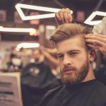 At the barber shop Handsome bearded man is reading a newspaper while getting haircut by hairdresser at the barbershop