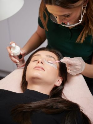 Woman having eyelash extension procedure in beauty salon. Close up of beauty specialist in sterile gloves applying cleansing mousse on woman eyelashes with cosmetic lash brush. Eyelash stylist preparing woman lashes for applying extensions.